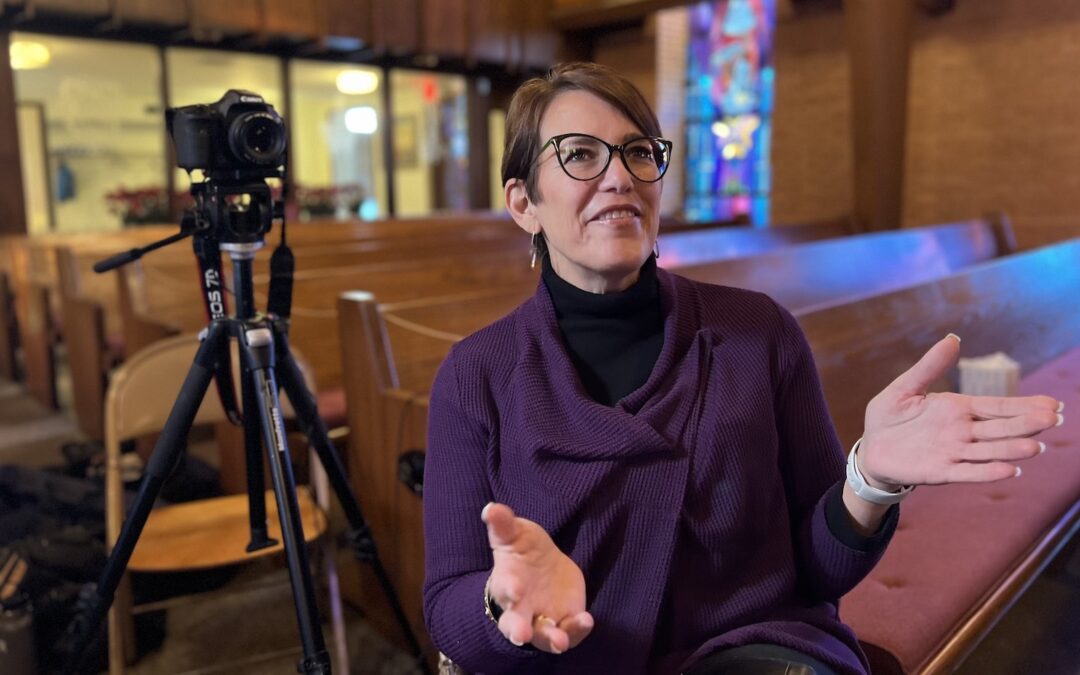 A woman sitting on a chair inside a church smiling and gesturing with her hands with a camera in the background.