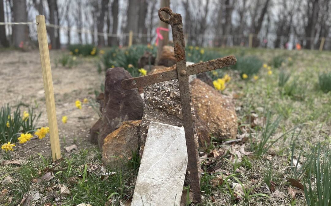 A rusty, metal cross in the ground next to a row of stones that serve as burial markers on the grounds of a Catholic church in Maryland.