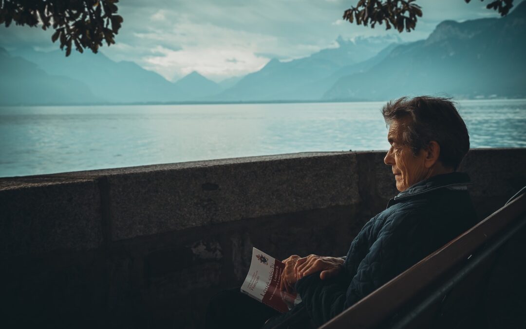 A man sitting outside on a bench holding a book in his lap and looking out at the ocean.