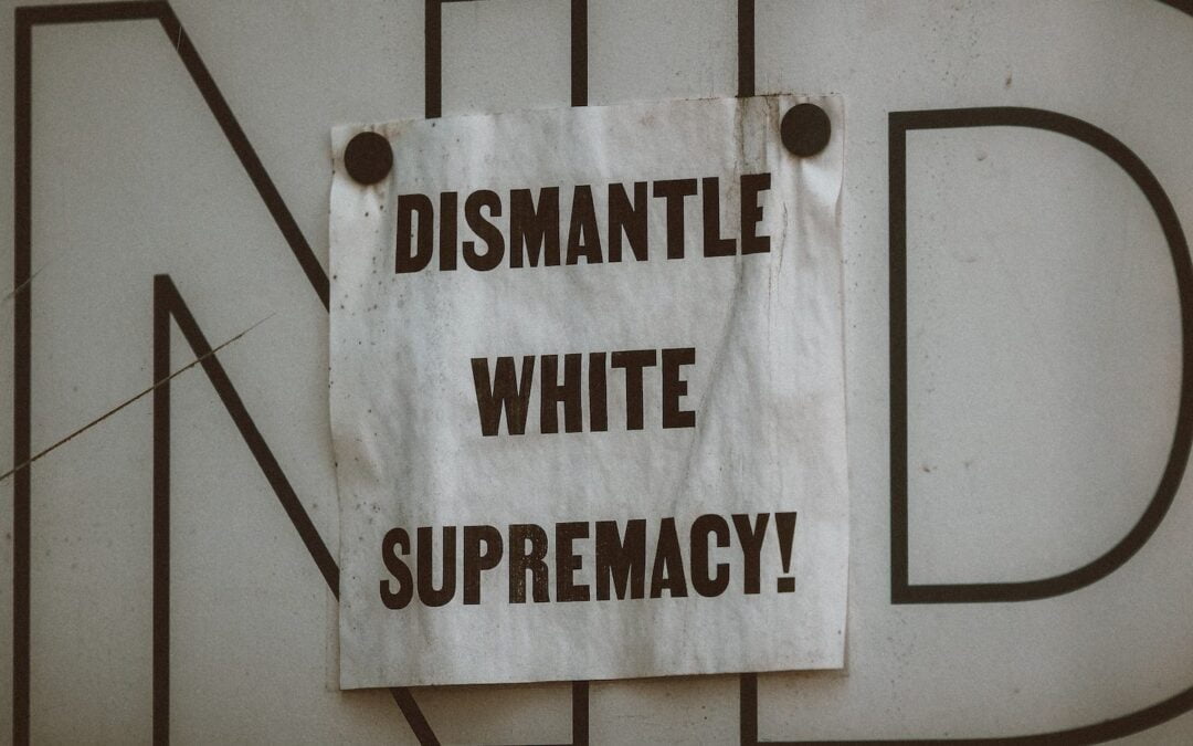 A white sign on a wall with bold, capital lettering that says, “Dismantle white supremacy!”