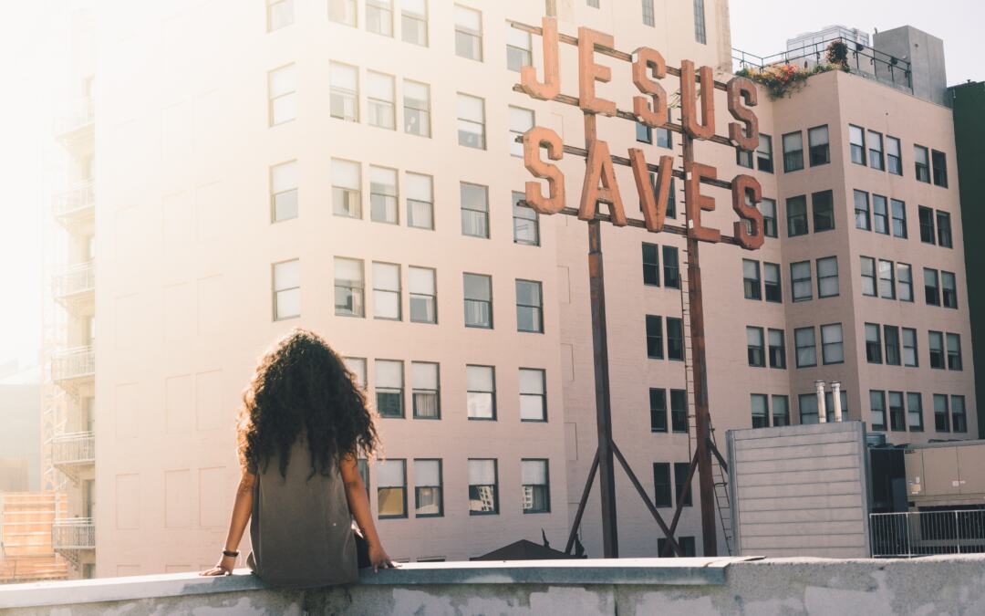 A woman sitting on the ledge of a building looking at a sign that reads, “Jesus saves.”