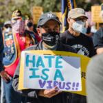 A man wearing a face mask and standing in a group of people holding a cardboard sign that says, “Hate is a virus.”