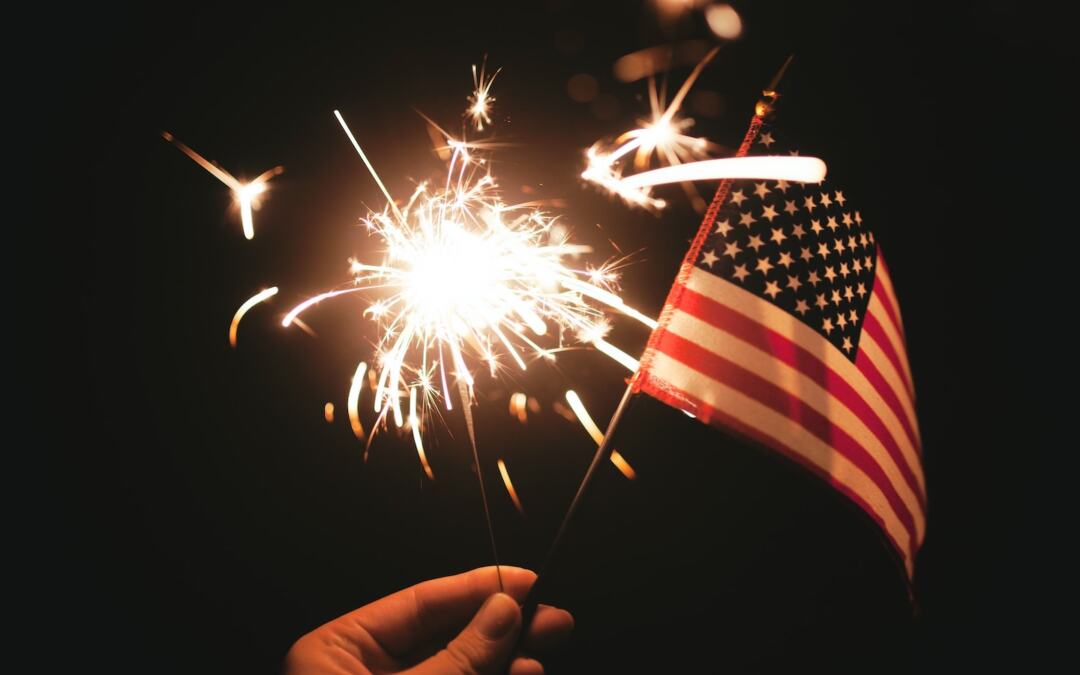 A person holding an American flag and a sparkler in one hand.