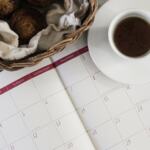 A table seen from above with a basket of muffins, a coffee mug and a calendar sitting on it.