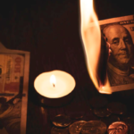 Several one hundred dollar bills on the ground, one burning near a candle.
