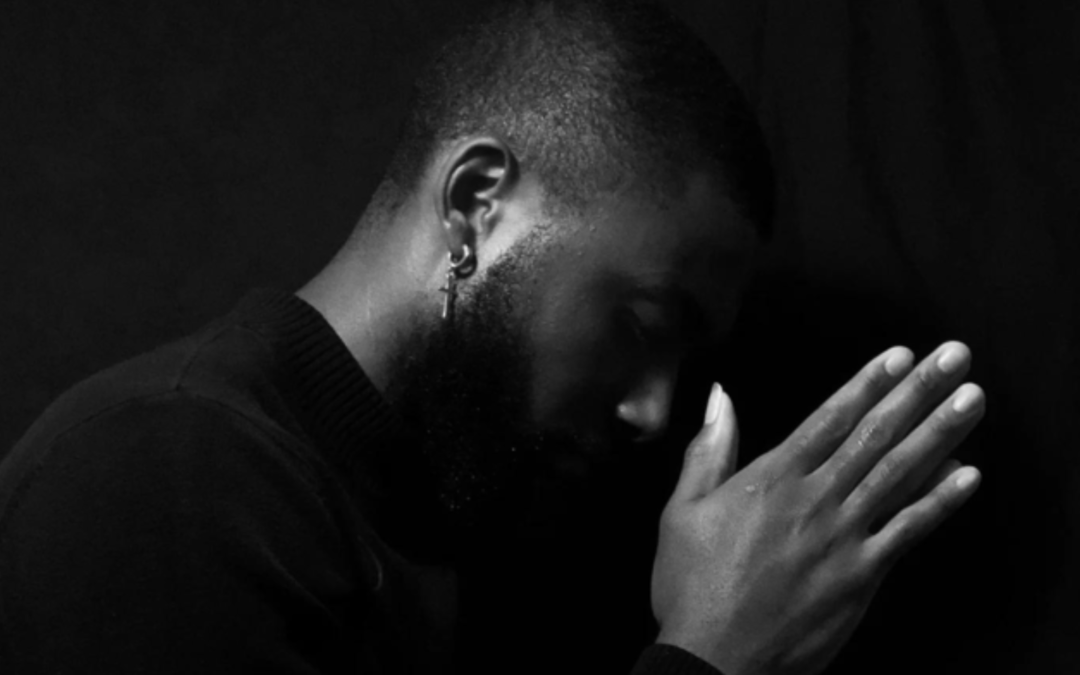 An African American with his hands pressed together and his head bowed against a black backdrop.
