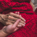 An elderly hospice patient in bed holding the hand of a loved one.