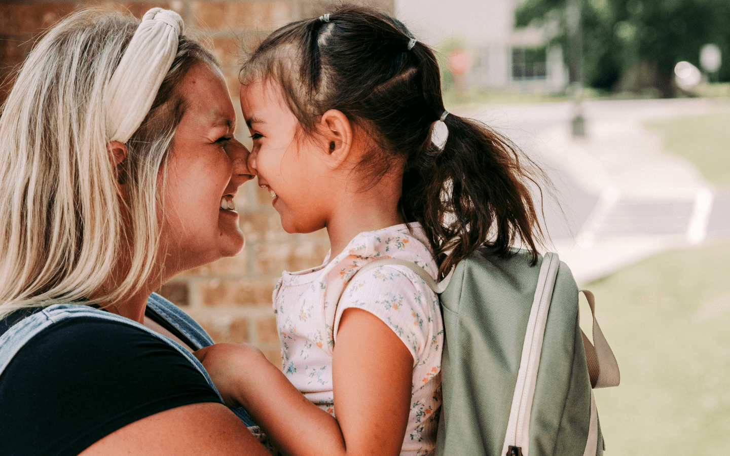 An adult greets a young child with a backpack at school.