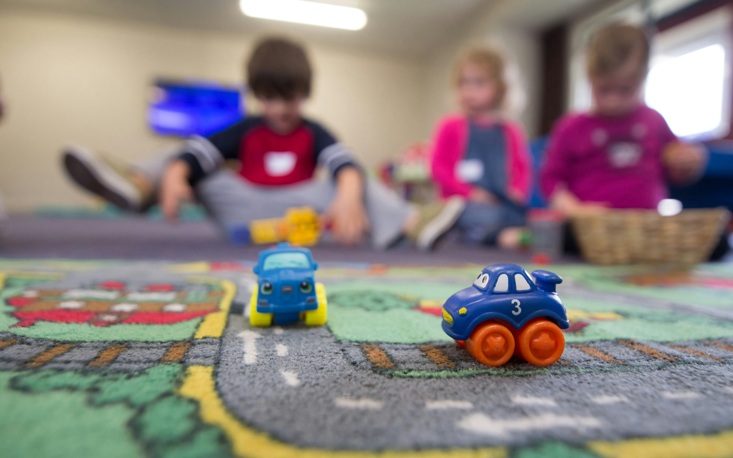 A photo of a daycare center with children playing on the floor and toy cars in the foreground.