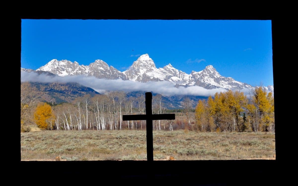 The Teton mountain range in Grand Teton National Park seen from a chapel with a cross in the center of the image.
