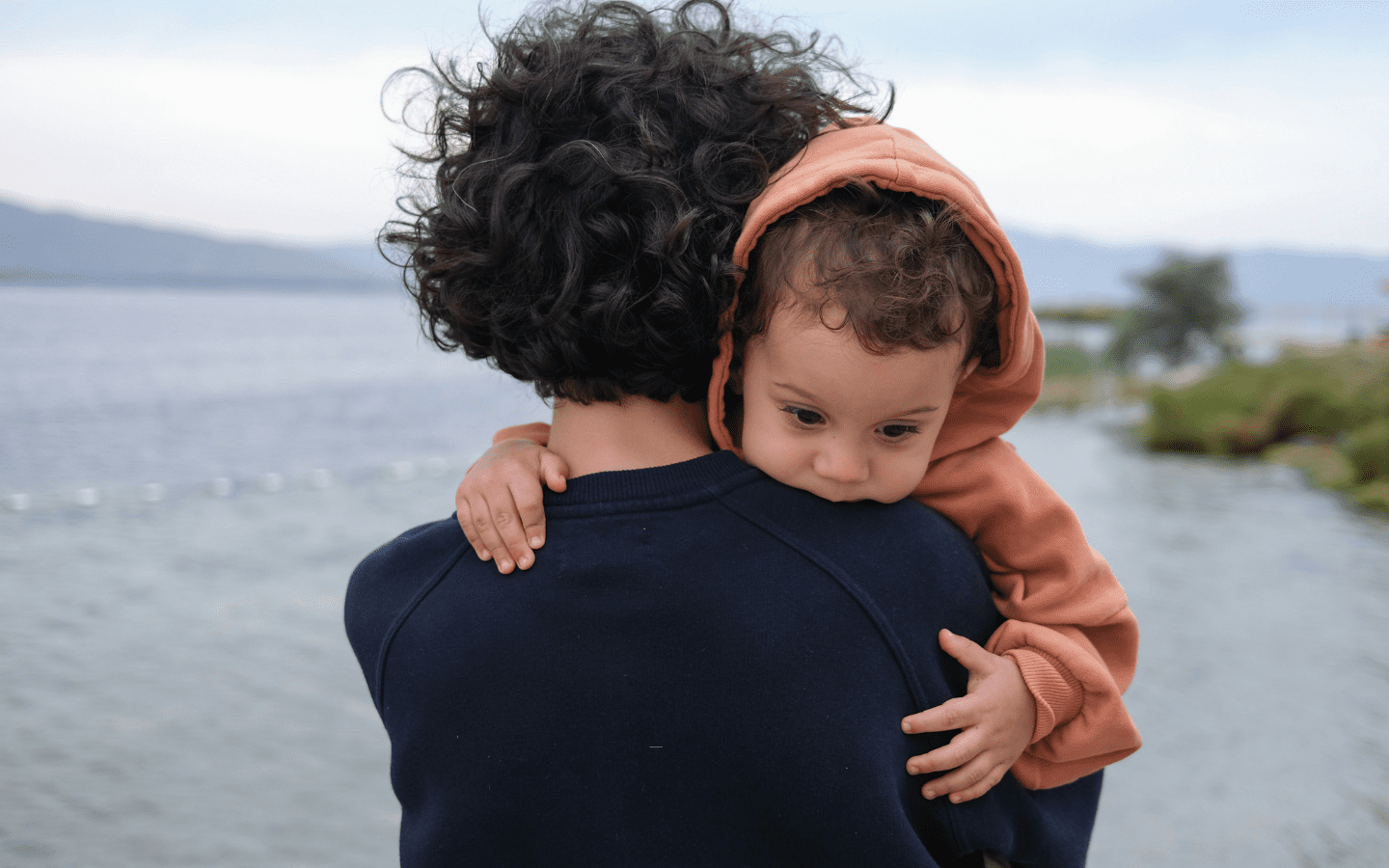 A young migrant parent looks across a body of water with their child.||