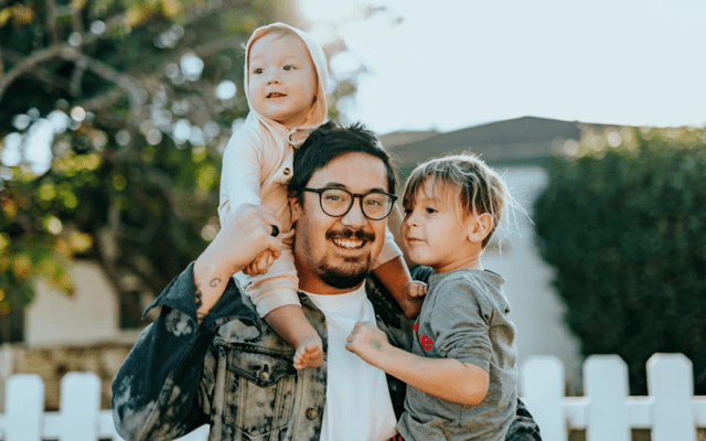 A man and his two children outside of a home with a white picket fence.