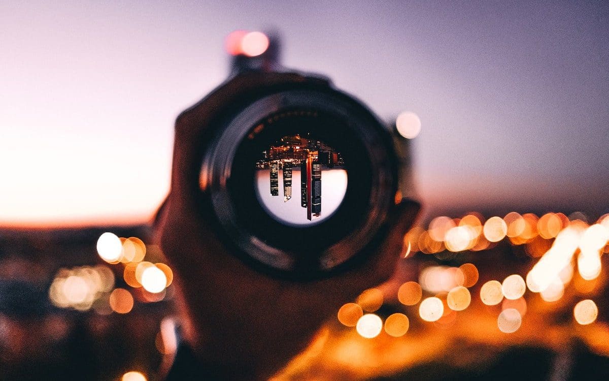 A person holding a camera lens through which a cityscape is seen upside down with the actual skyline blurred in the background.