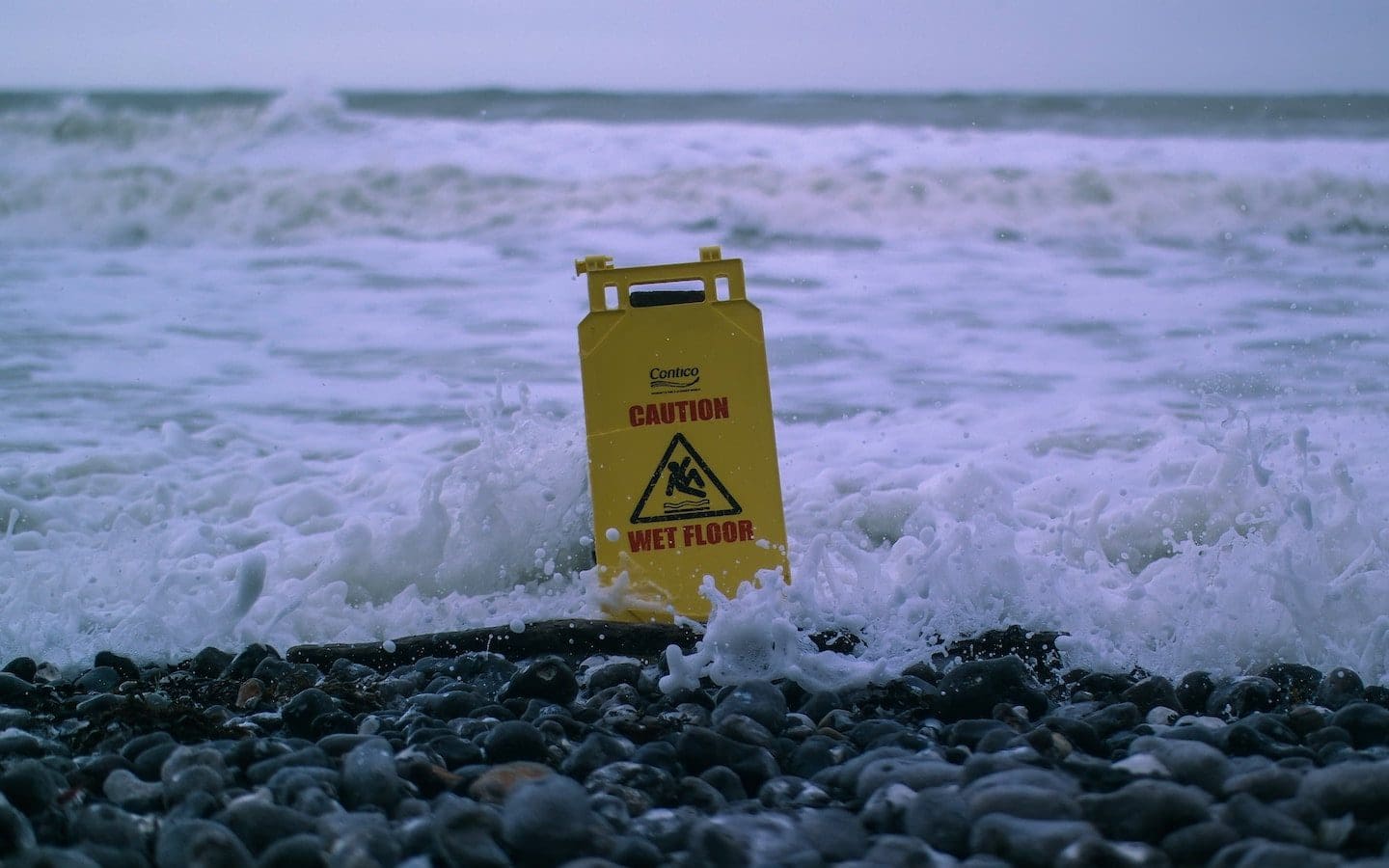 A yellow caution wet floor sign sitting in the surf at the edge of a rocky shoreline.