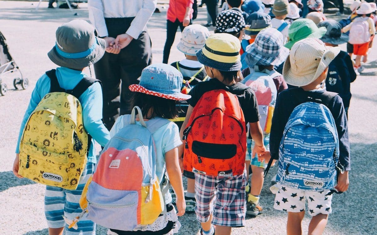 A row of school-age children seen from behind walking and wearing backpacks.