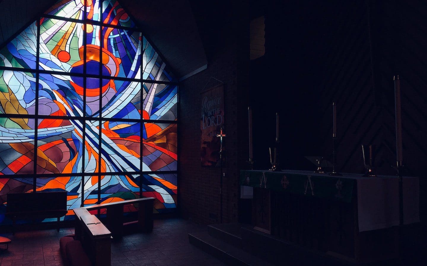 The interior of a church worship space with the room dark except for light shining through a stained-glass window.