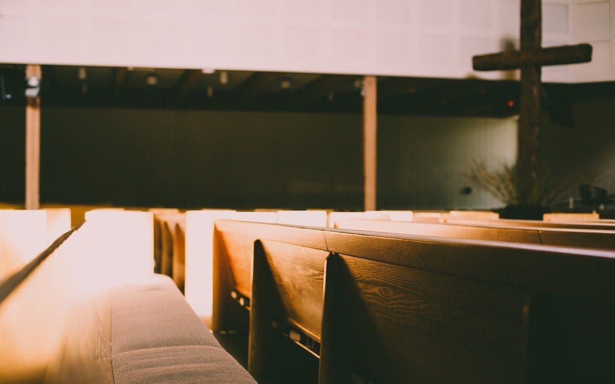 The interior of a church sanctuary with empty pews in the foreground and a wooden cross in the background.
