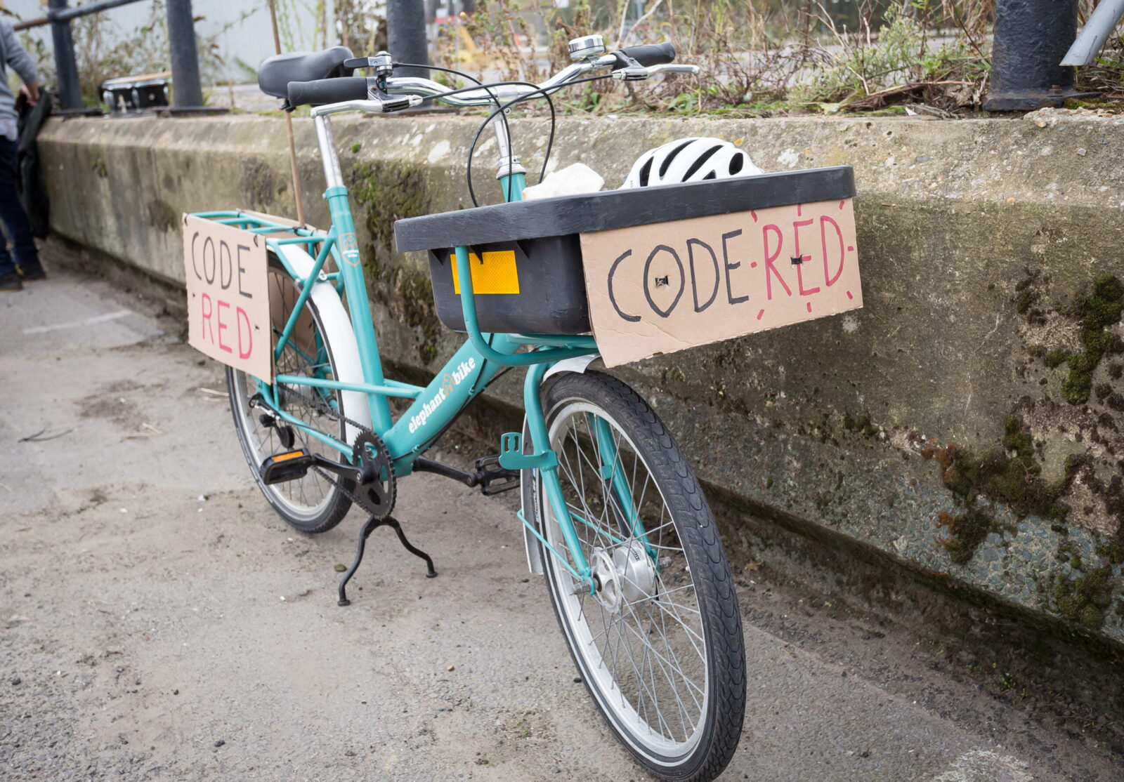 A bike parked on a sidewalk with the words “code red” on cardboard signs attached to the front and rear of the bike.