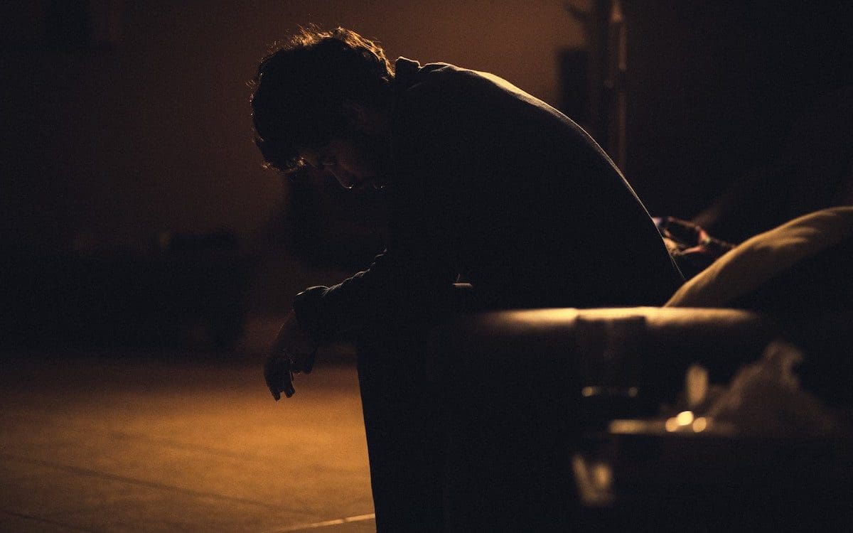 A man sitting on a couch in a darkened room with arms on knees and head bowed.