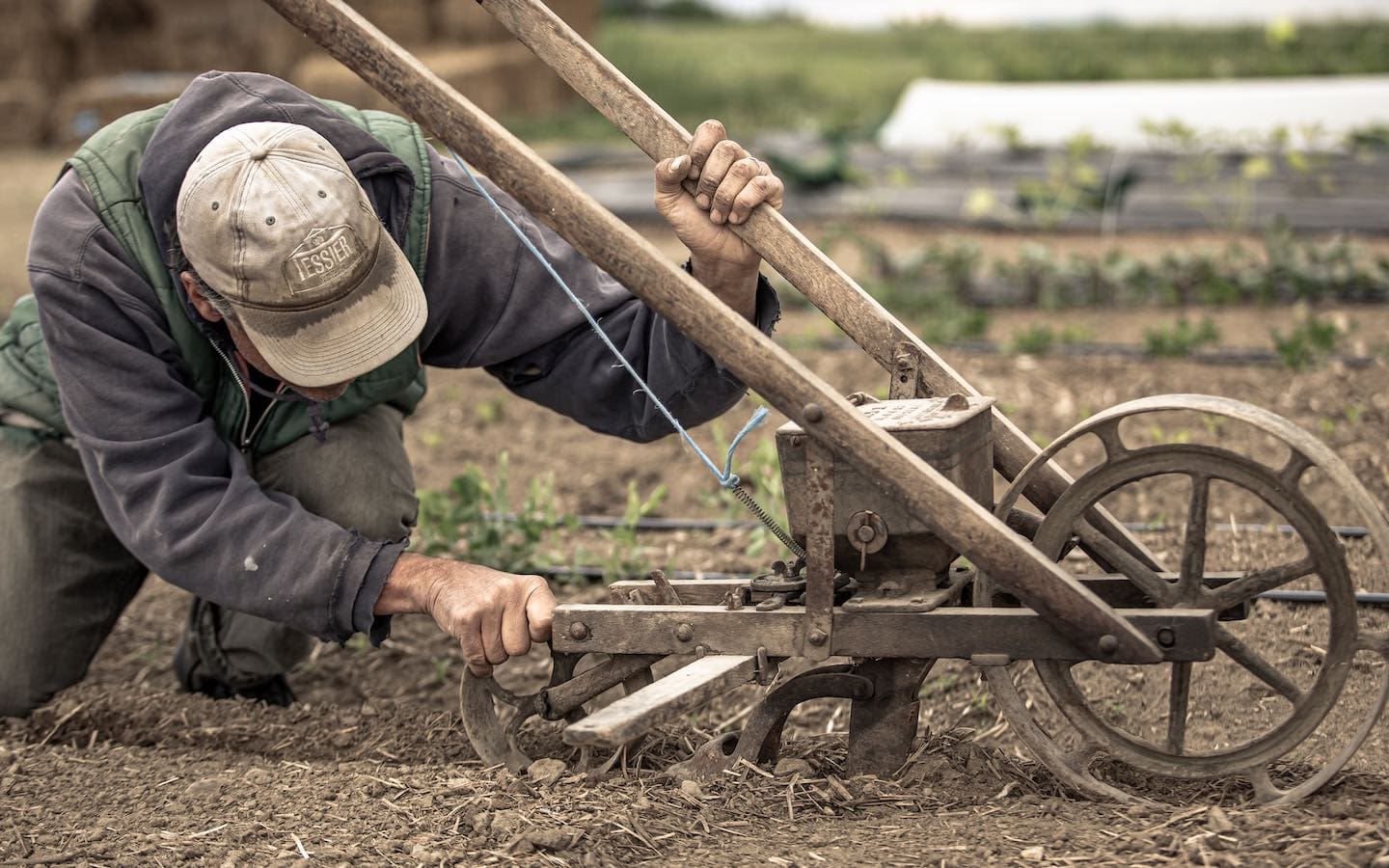 A farmer kneeling on the ground in a field next to an old-style plow to make adjustments.