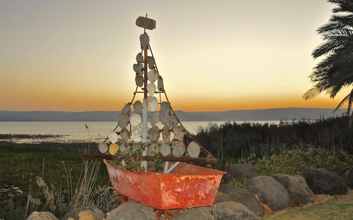A model boat with rock carvings as the sails with a sunrise over the Sea of Galilee in the background.