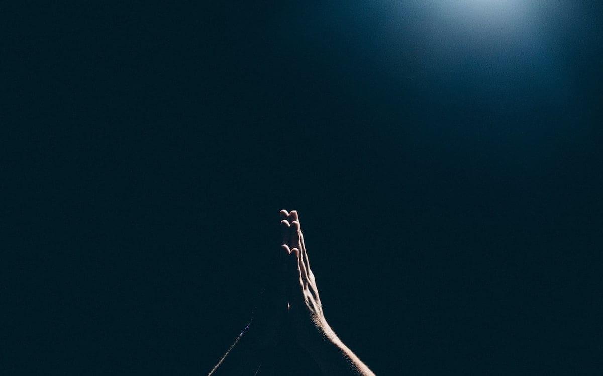 Hands put together to indicate praying with a slight light in the upper right and the rest of the image in darkness.