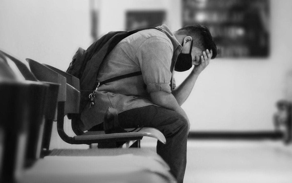 A man in a black jacket sitting on a bench with his head in his hands.