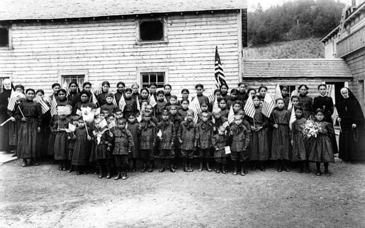 Indigenous children in school uniforms holding American flags in front of a mission school in Holy Cross