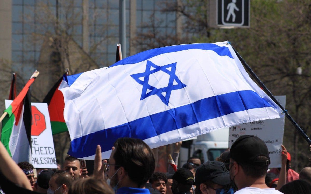 An Israeli flag in the center of the image with a Palestinian flag in the background to the left.