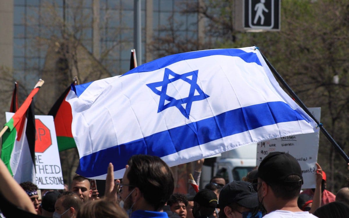 An Israeli flag in the center of the image with a Palestinian flag in the background to the left.