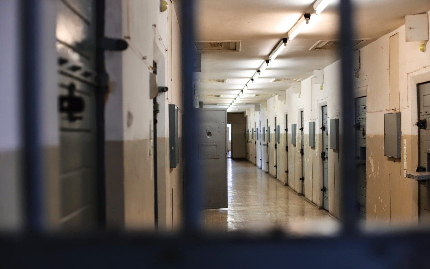A jail or prison cell seen from the inside looking past the bars to a hallway with other cells.