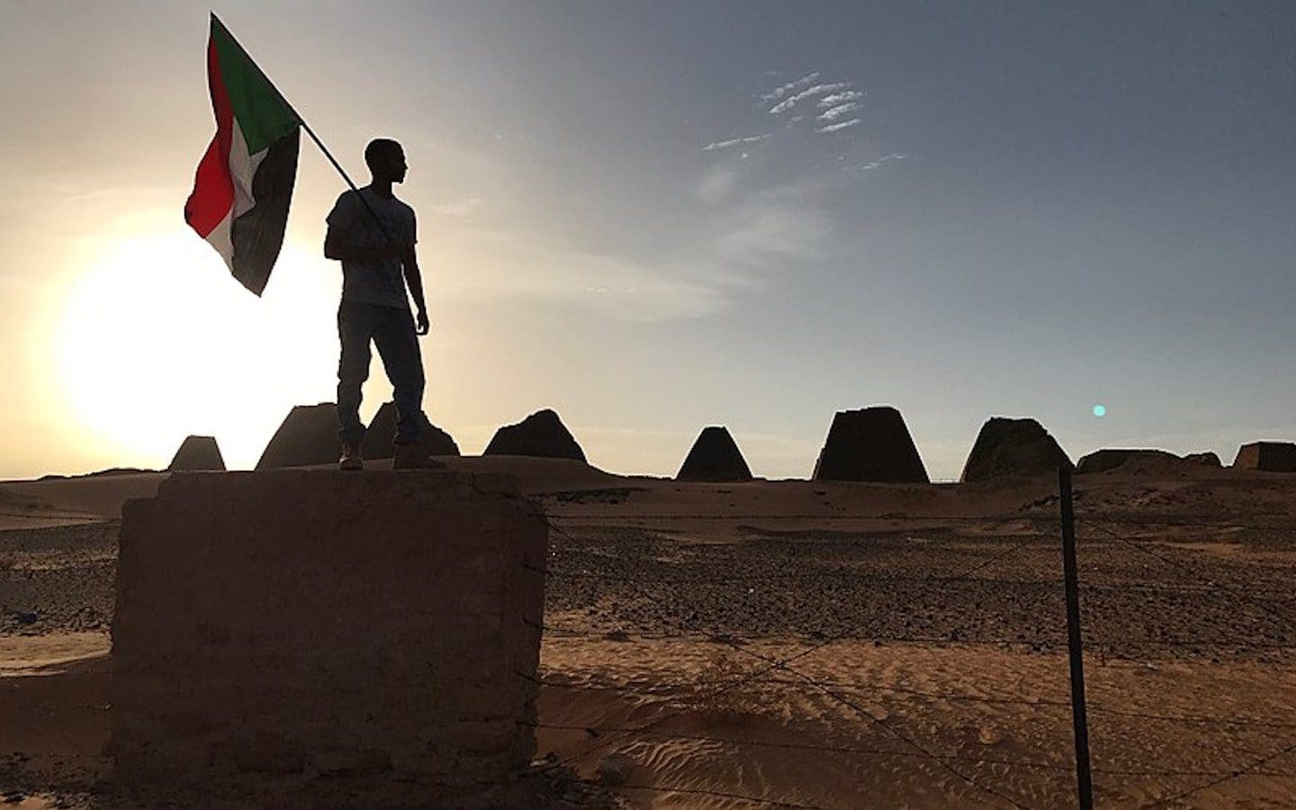 The silhouette of a man holding a Sudanese flag standing on a concrete wall with the sun setting in the background.