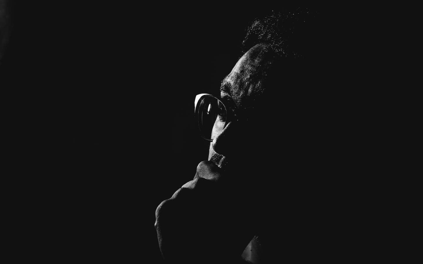 A man sitting in a dark room with his hand resting on his chin in a posture of thinking / reflecting / contemplating.