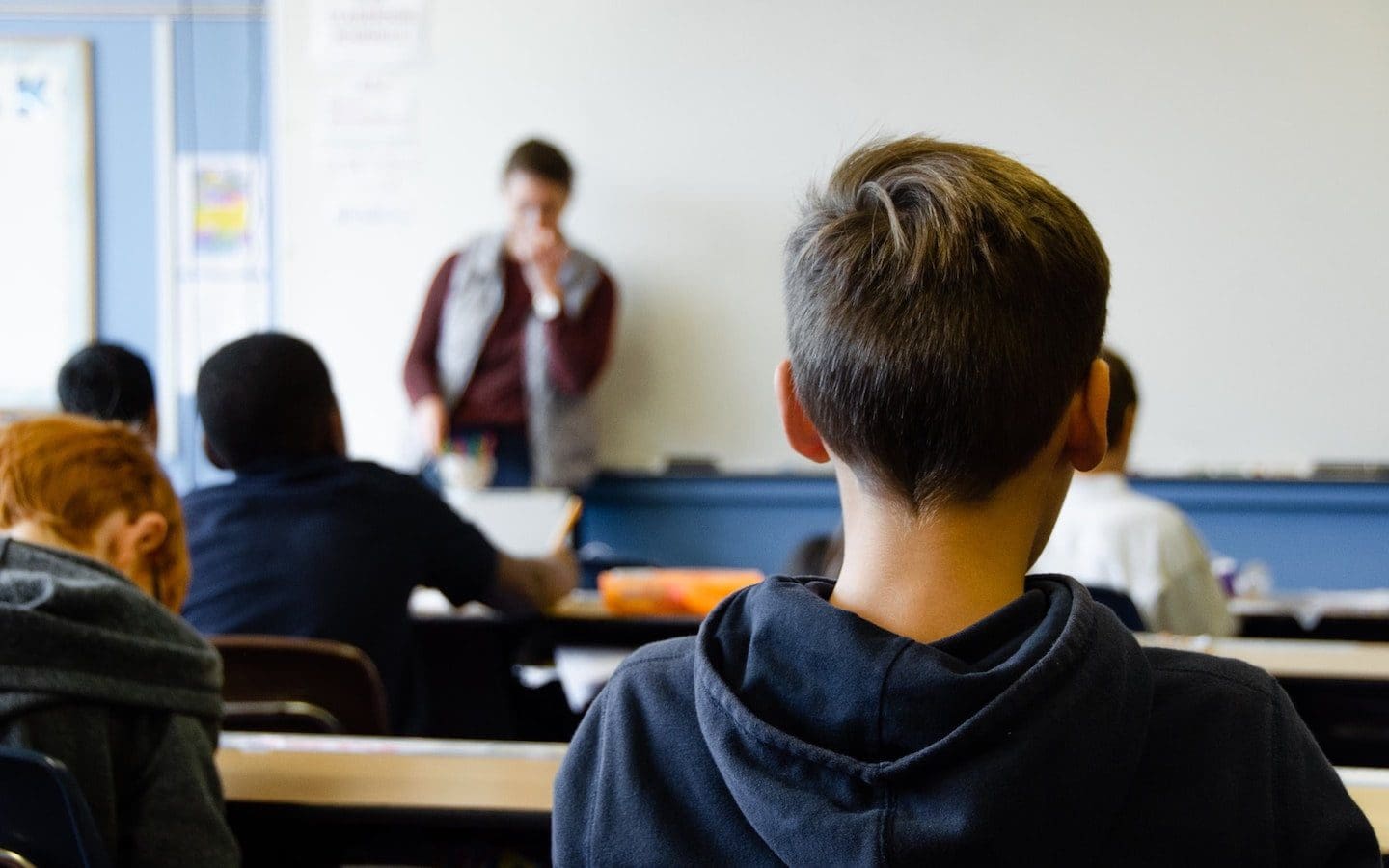 Students sitting at desks in a classroom seen from behind with a teacher standing at the front by a whiteboard.