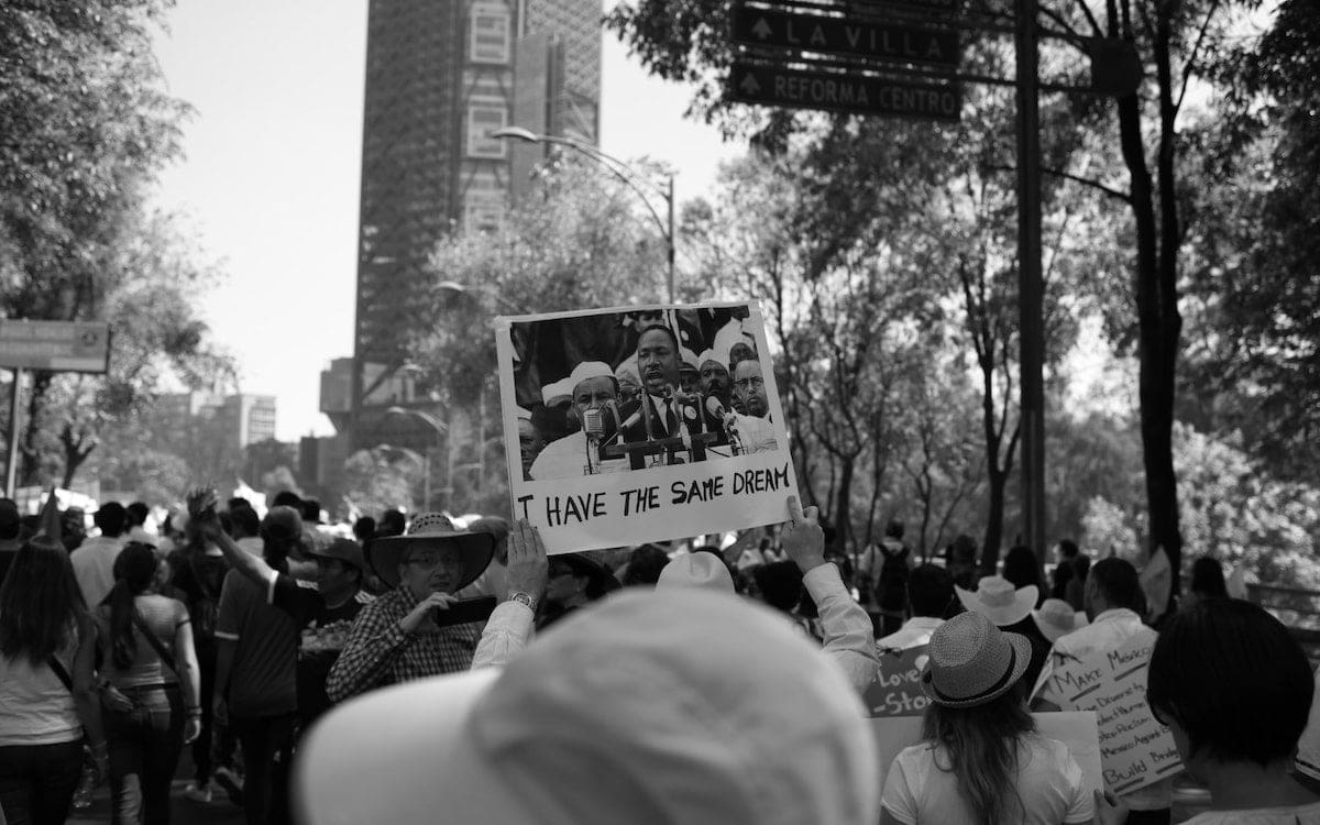 A crowd on the streets seen from behind with a person holding up a sign with a photo of Martin Luther King Jr speaking that says