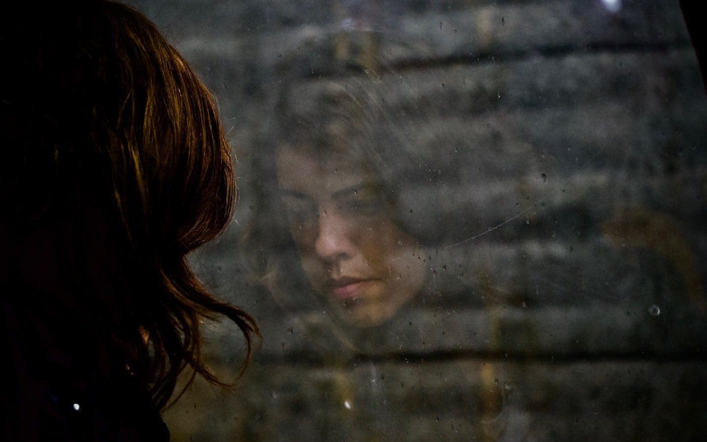 A woman seen from behind looking out a window with her face reflected in the window.