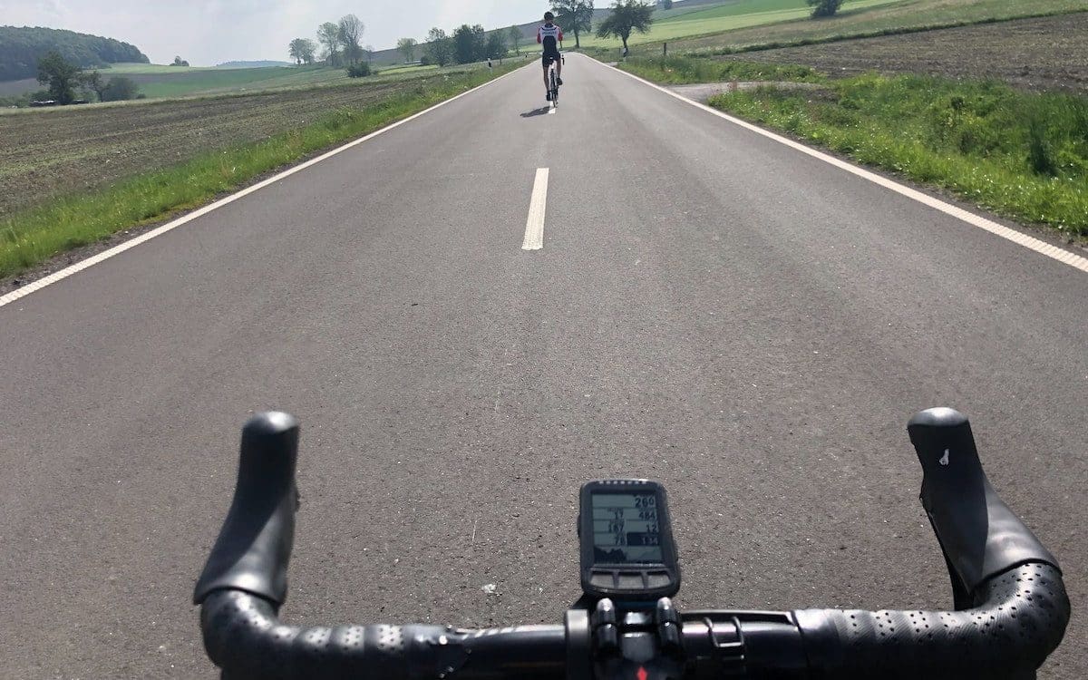 A road with a biker in the distance and a bike’s handlebars at the bottom of the image.|A man in a biking shirt holding up a race medal.