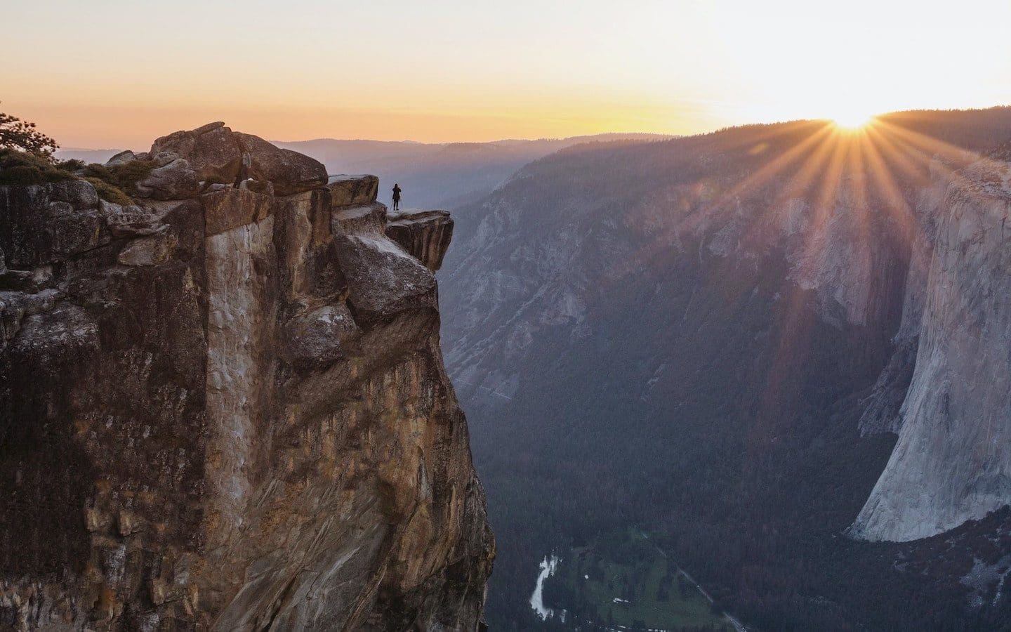 A rocky cliff with a person standing at the edge looking at the sun rising over another cliff in the distance.