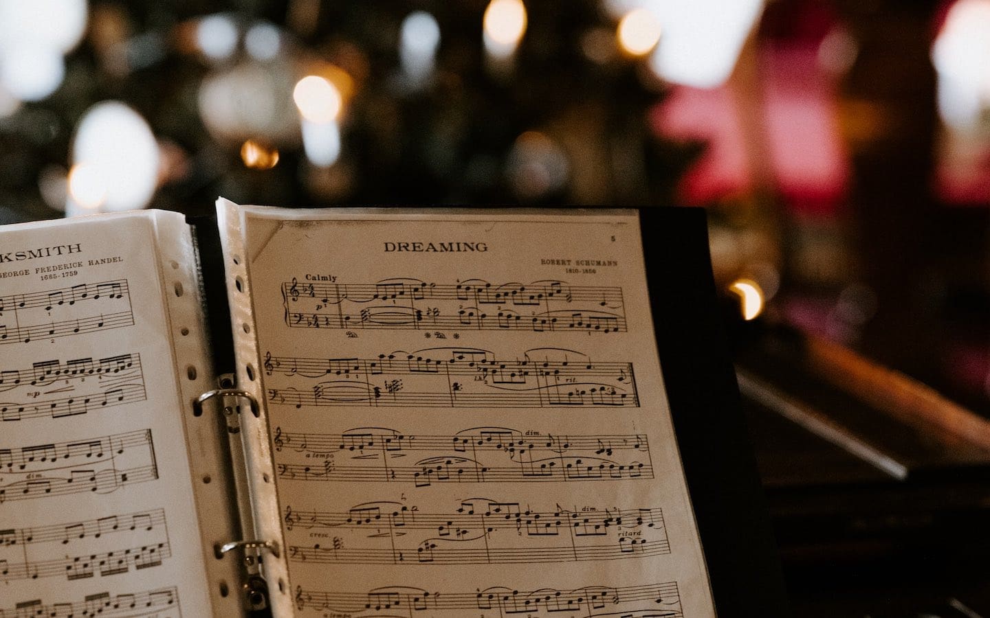 A piano with sheet music on it in the foreground and a lighted Christmas tree in the background.