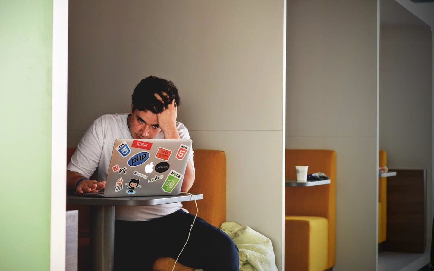 A college students sitting at a table looking at his computer with his hand on his head and a stressed look on his face.