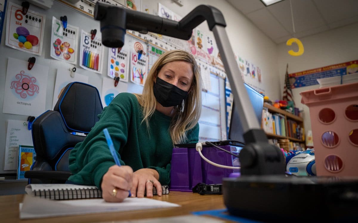 A teacher wearing a mask while sitting at her desk writing.