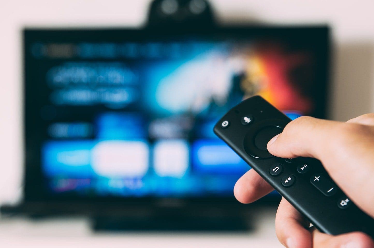 A remote control in focus in the foreground and a TV out of focus in the background.
