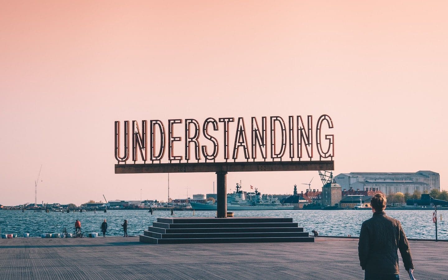 A metal sign in block lettering that says “Understanding” on a boardwalk near water.