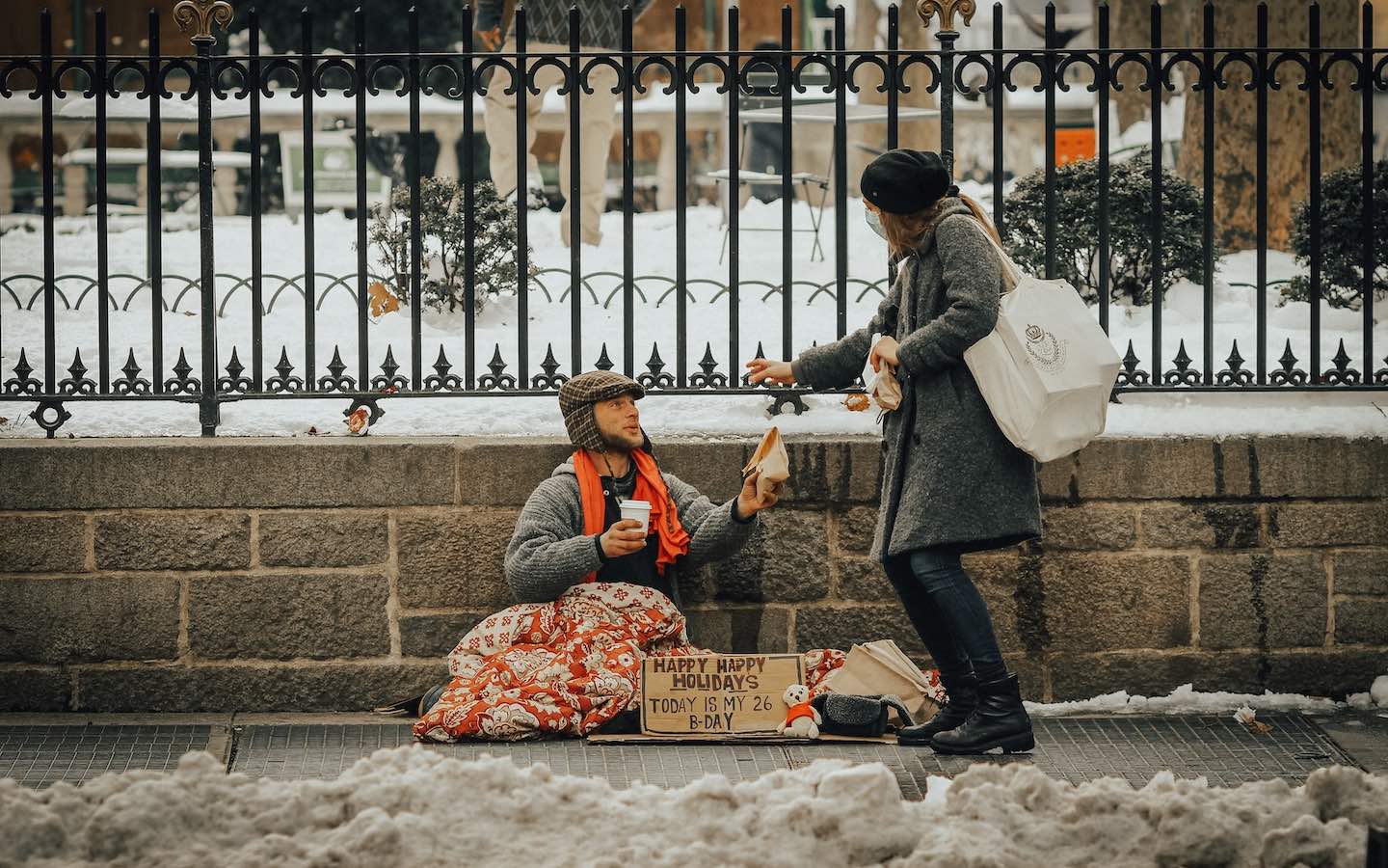 An unhoused person sitting on a street during the winter with someone handing them some food.