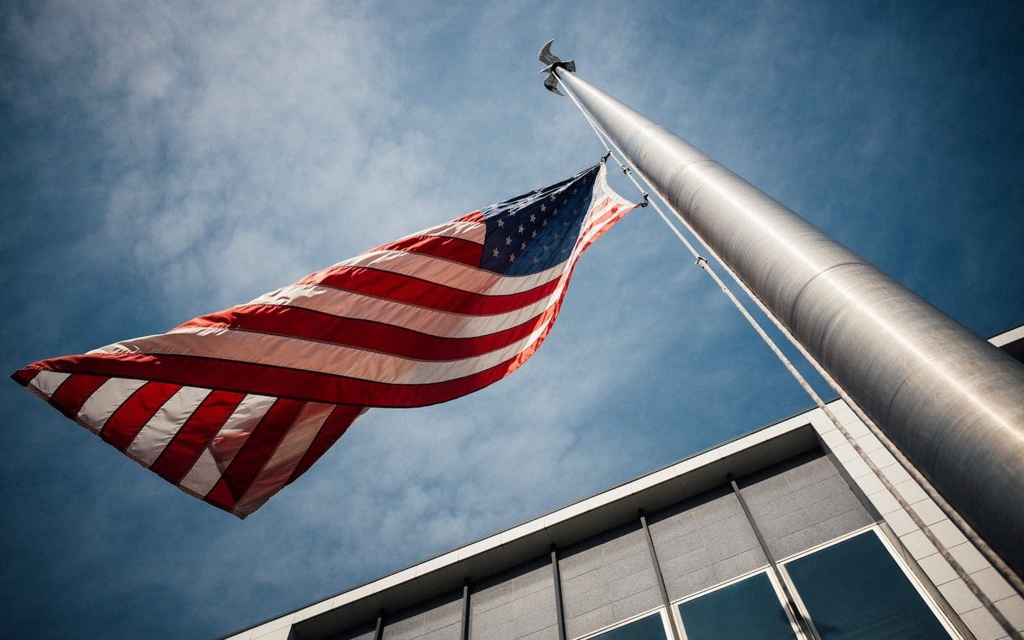 A U.S. flag at half-mast seen from below with a grey building in the background and a cloudy sky.