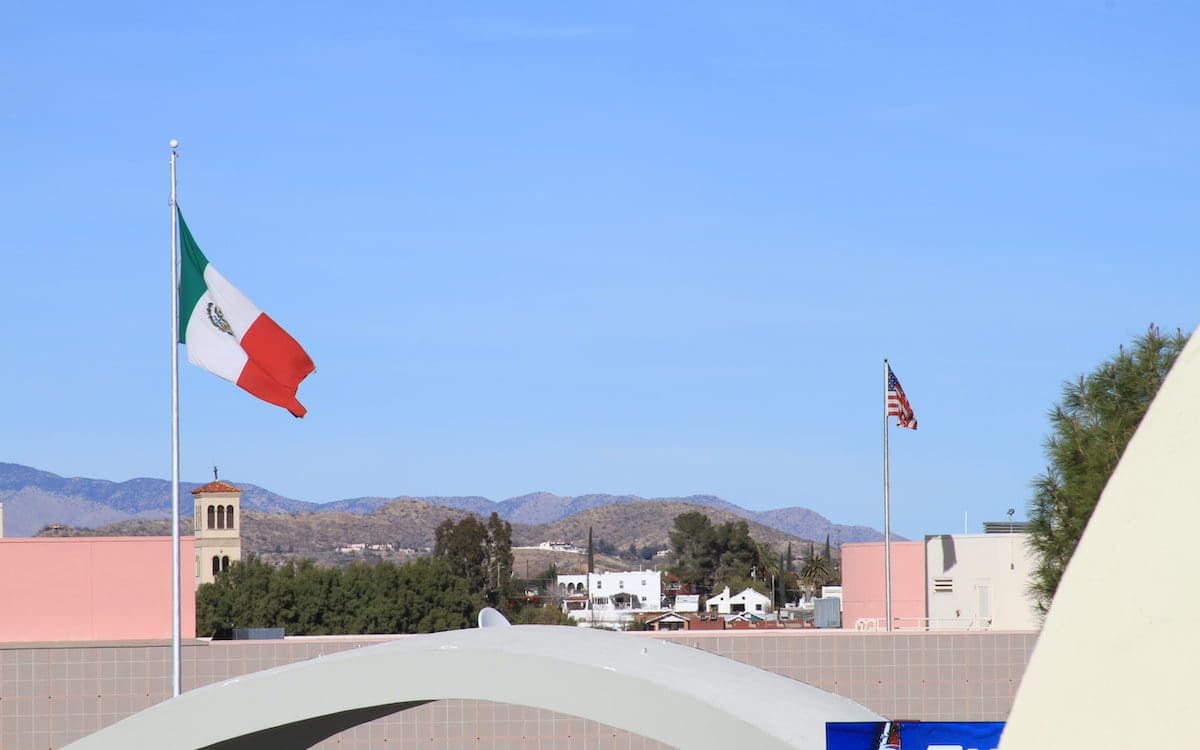 A Mexican flag in the foreground and a U.S. flag in the background along the U.S.-Mexico border.