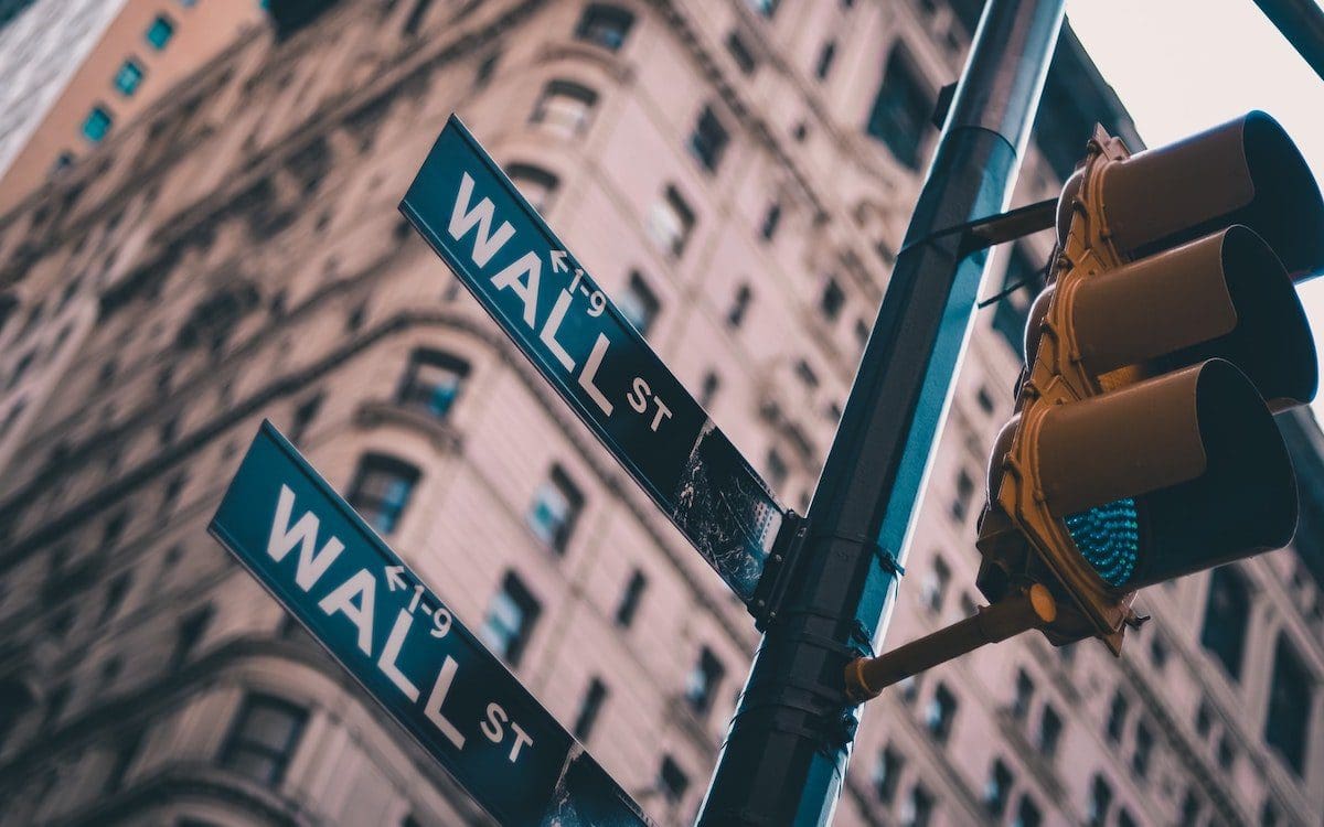 Looking up at street signs on the corner of Wall Street in New York City with buildings seen from below in the background.