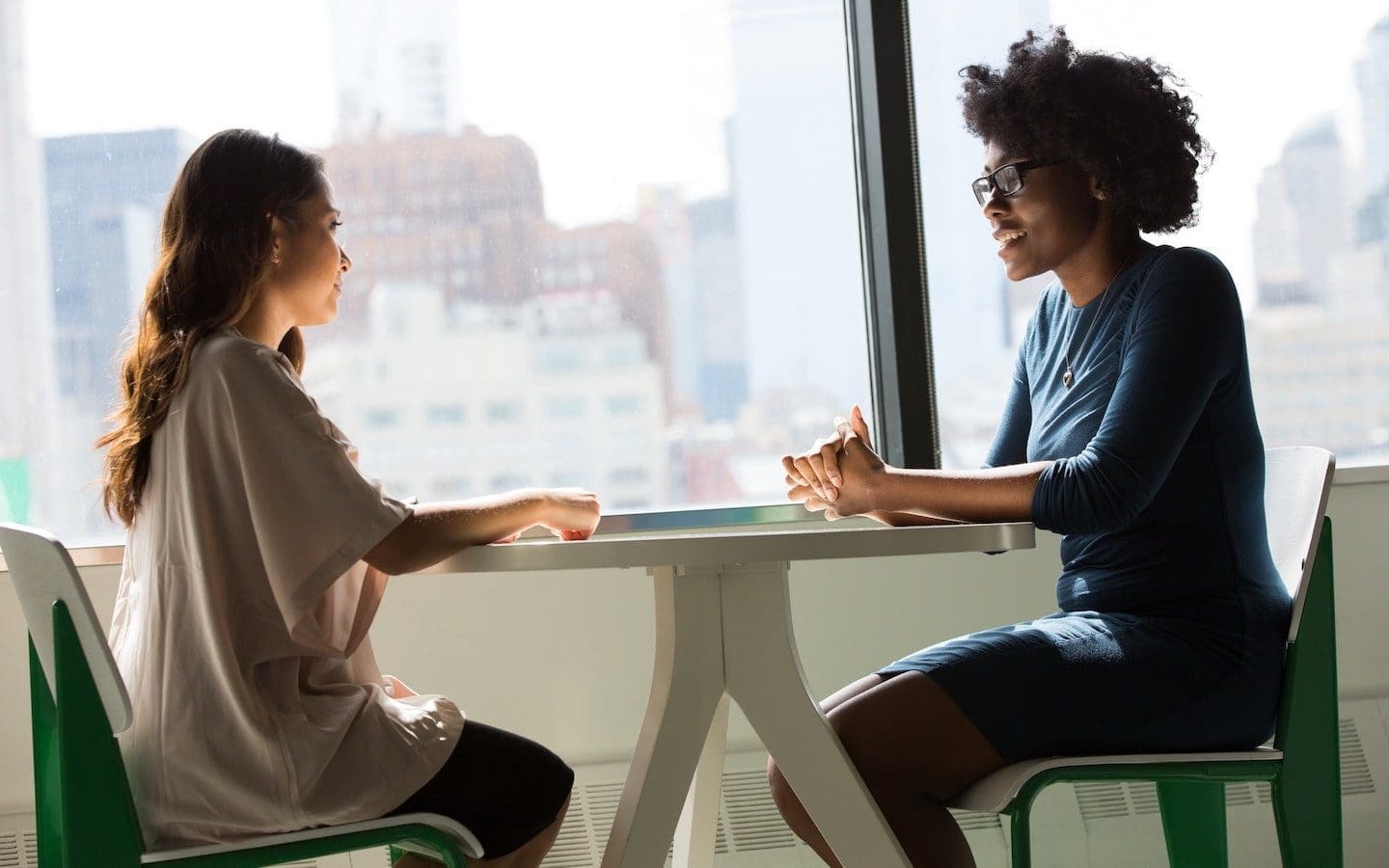 Two women sitting at a table with a large window revealing a city skyline in the background.