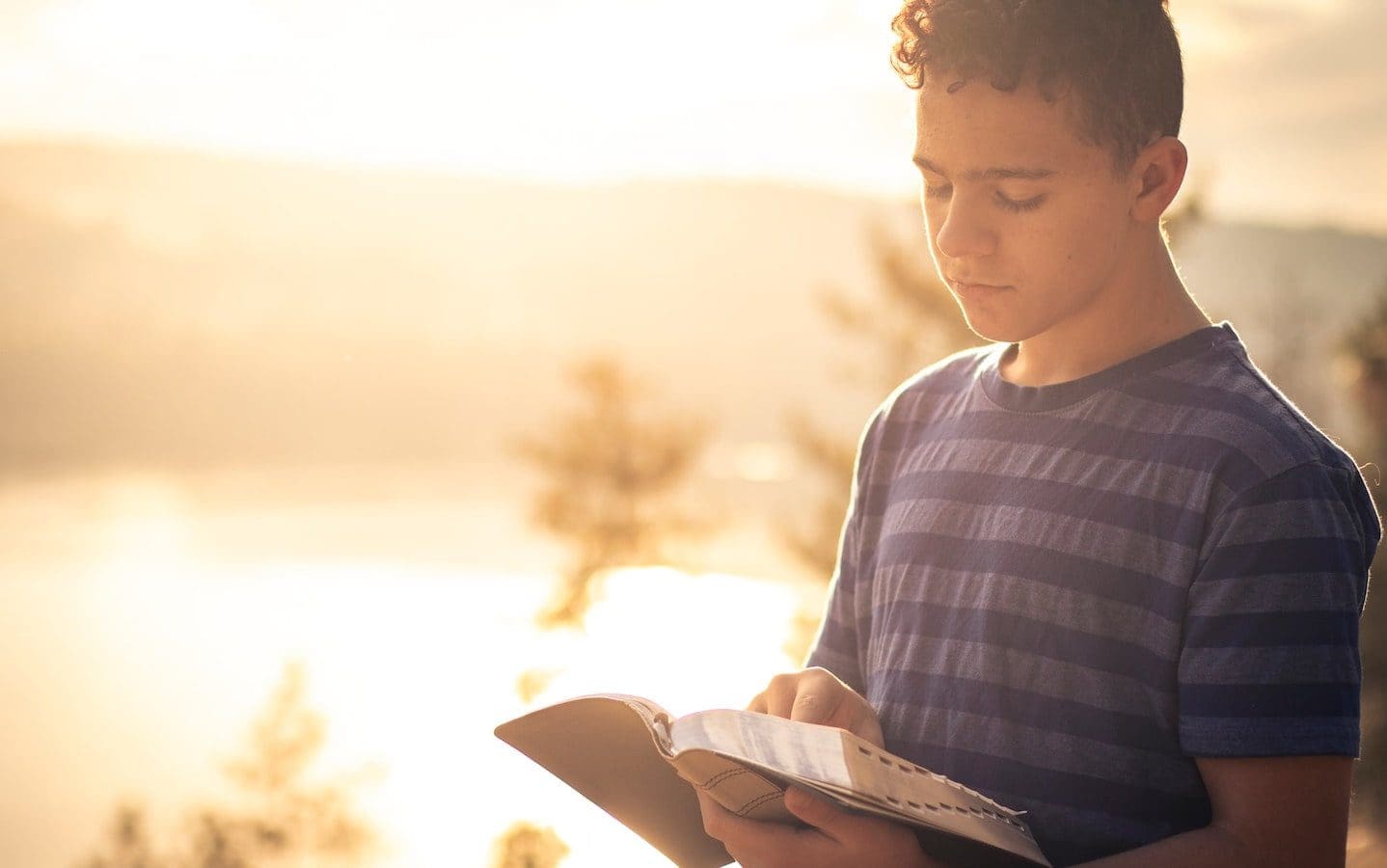A young adult standing outside holding an open Bible that he is reading.