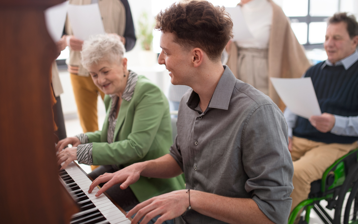 A young man and older lady play the piano together at a senior living facility.